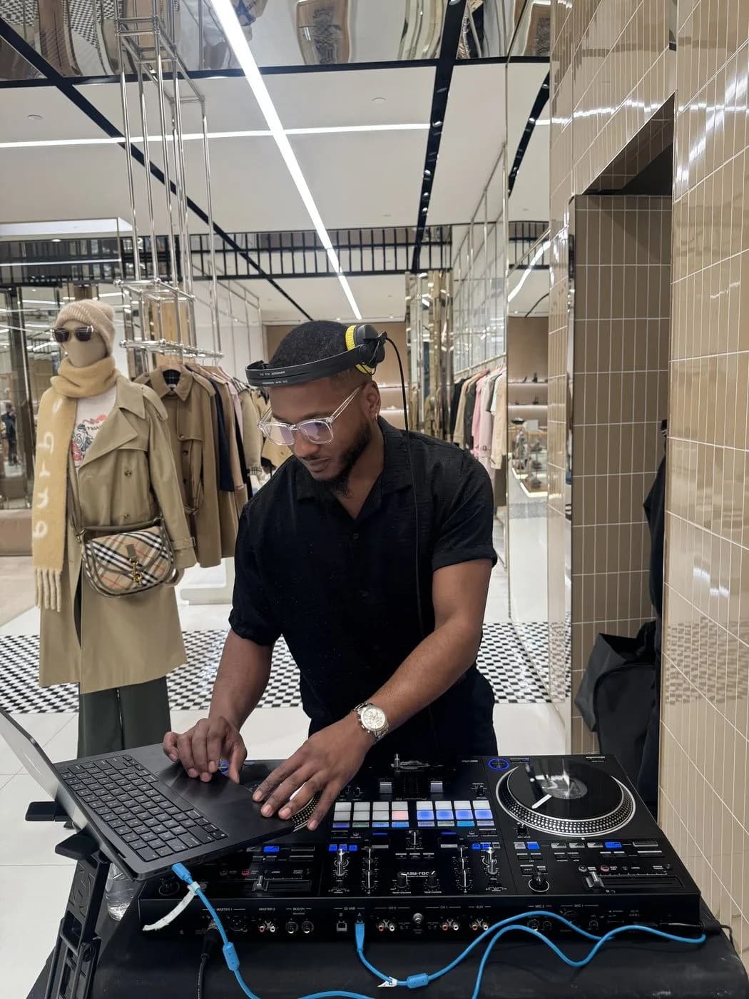 DJ Trizzy mixing on a controller inside a Burberry flagship store, beside a mannequin in a Burberry trench coat