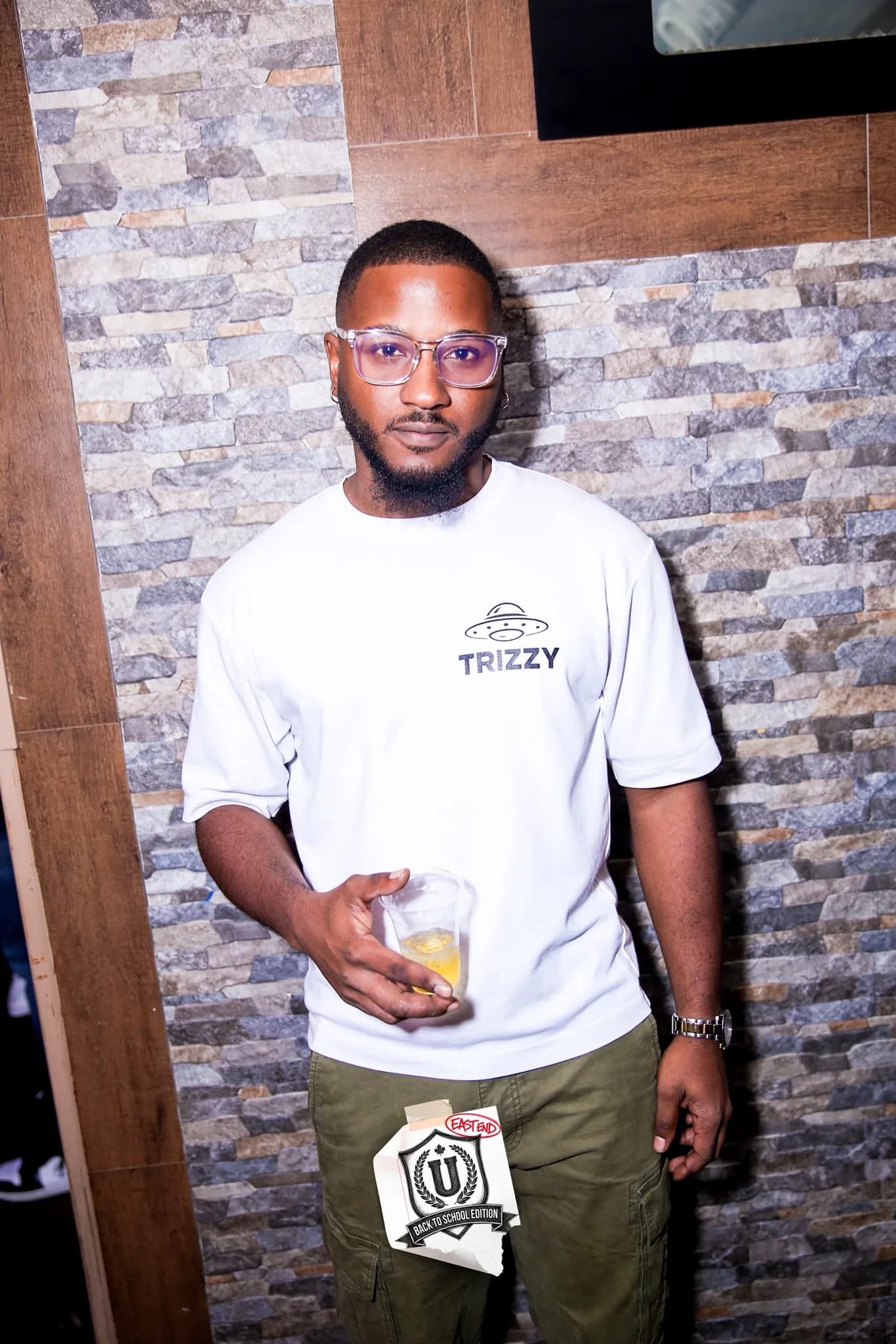 DJ Trizzy in a white TRIZZY UFO-logo tee and clear glasses, drink in hand, against a stone wall